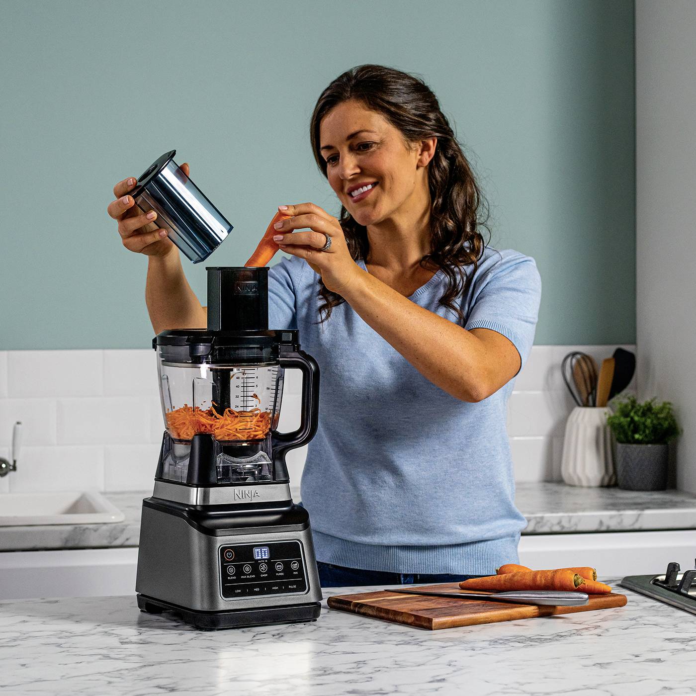 Een vrouw gebruikt een staande blender in een keuken. De blender bevat geraspte wortels. Op het aanrecht ligt een snijplank met wortels.
