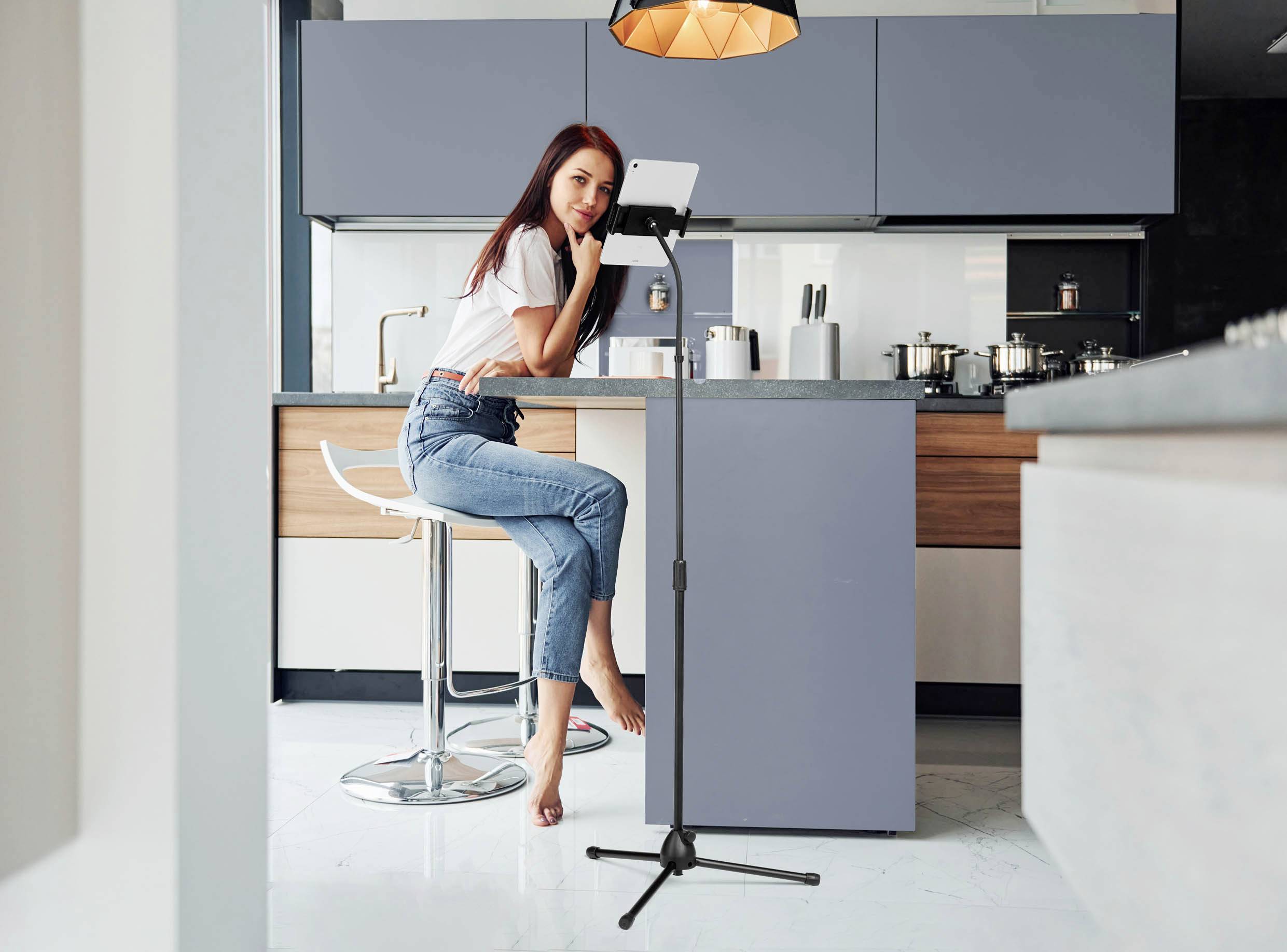 Een vrouw zit op een barkruk in een moderne keuken en kijkt naar een tablet dat op een standaard is bevestigd. Achtergrond met keukenmeubilair.