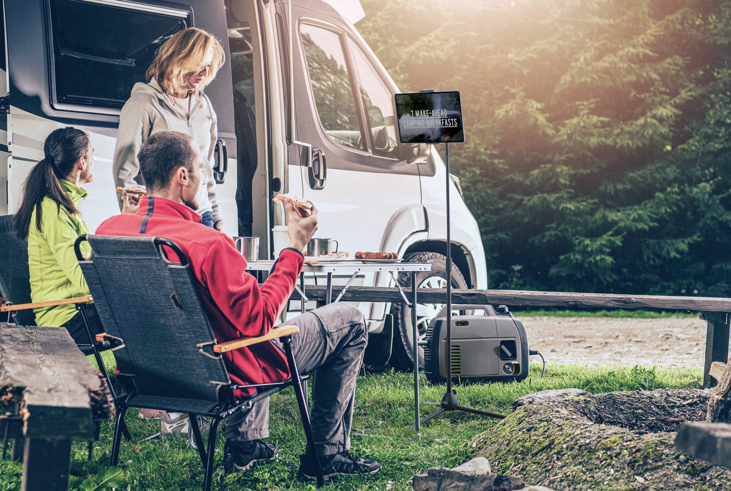 'Mensen ontspannen bij het kamperen voor een camper. Twee zitten op stoelen te eten, terwijl een persoon staat. Bos op de achtergrond.'