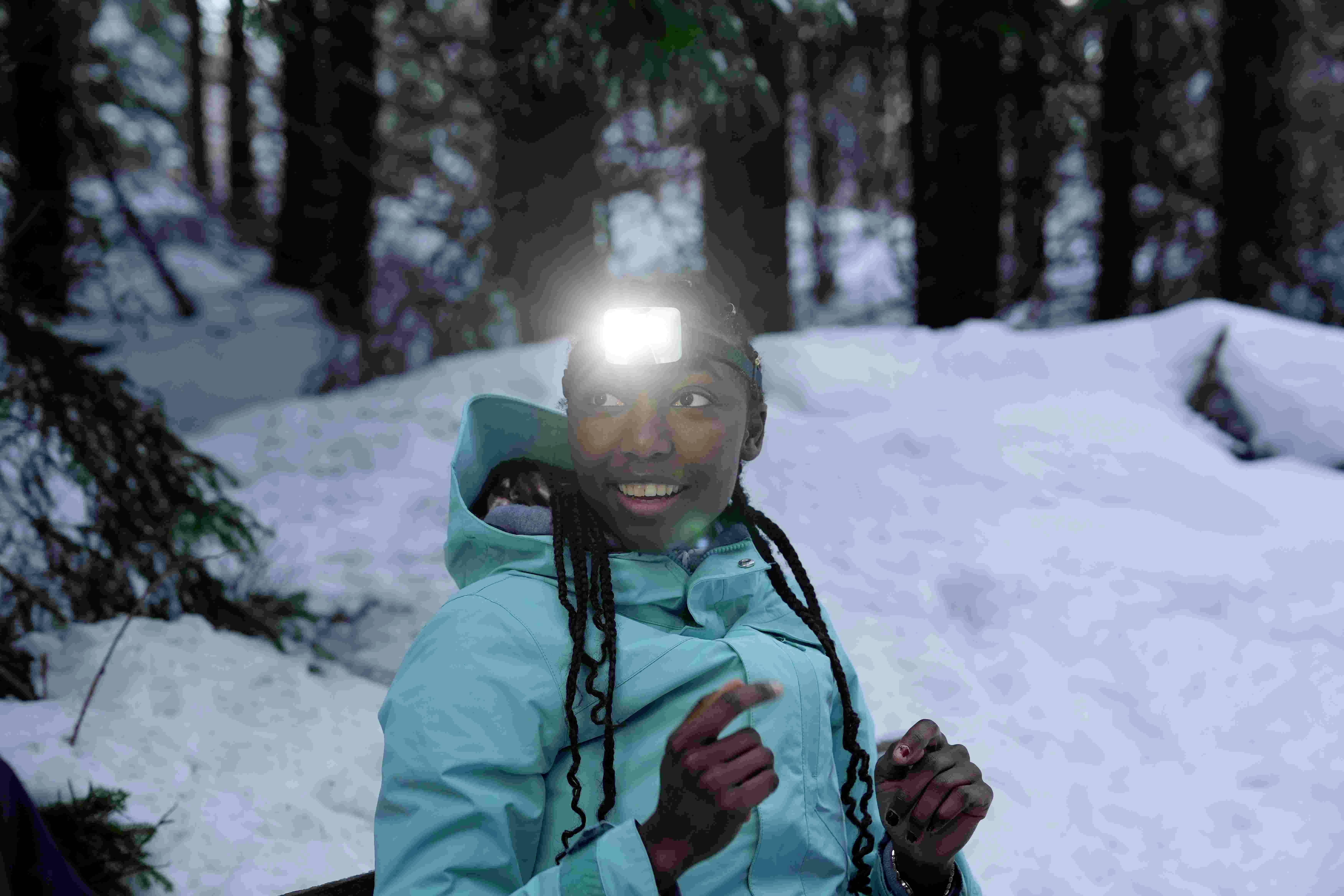 Persoon in een winterjas met een hoofdlamp in het besneeuwde bos, glimlachend en naar links kijkend.