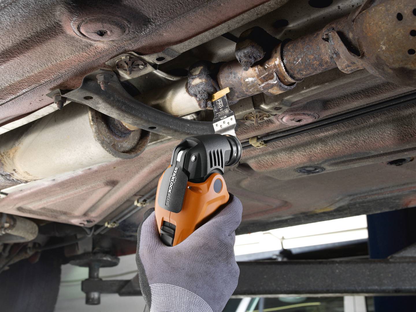 Een monteur gebruikt een oranje elektrisch gereedschap om een verroeste verbinding van auto-onderdelen onder een voertuig te bewerken.