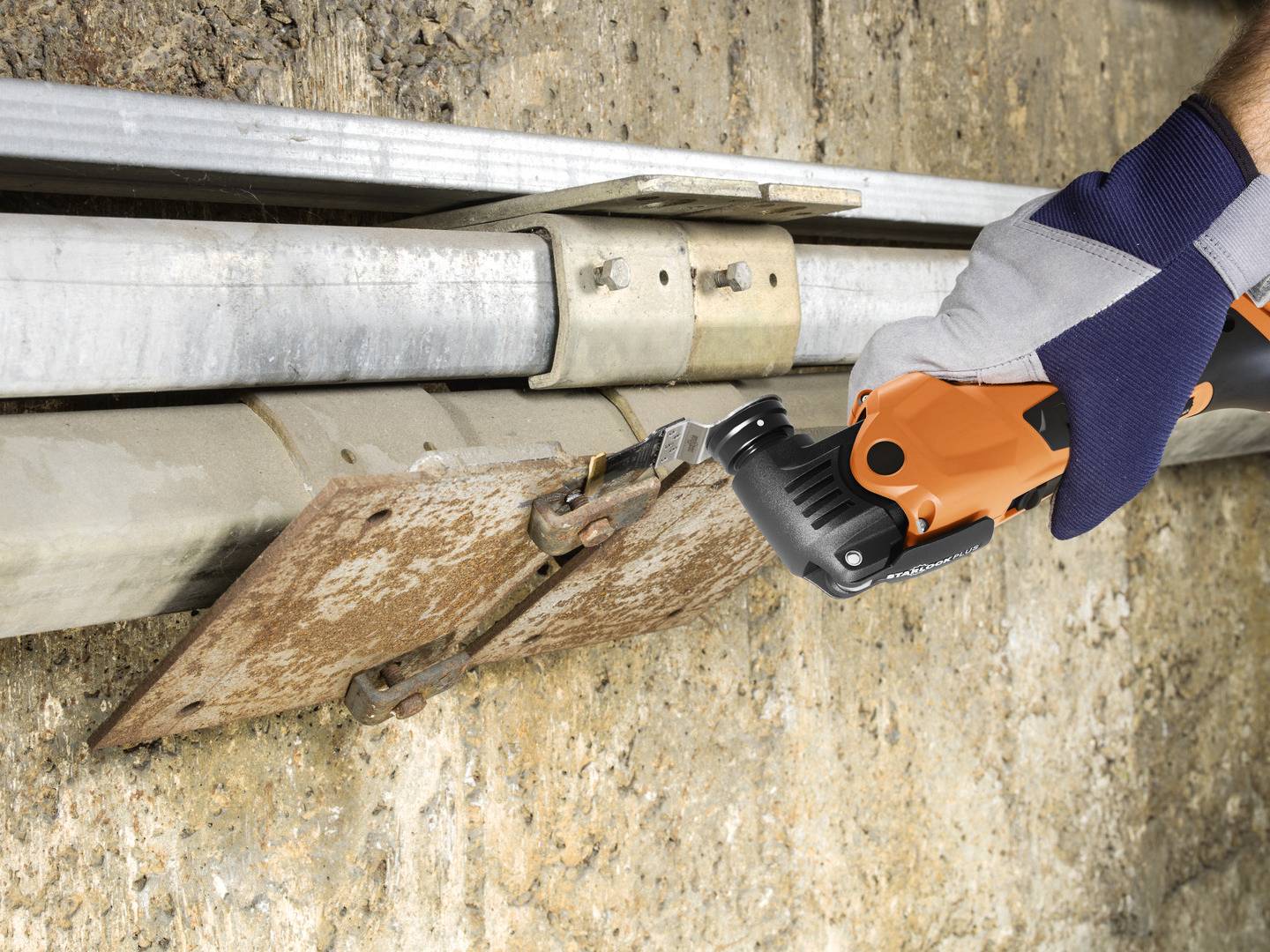 Een hand houdt een oranje elektrisch gereedschap met een zwarte handgreep vast, dat werkt aan een roestig metalen scharnier op een betonblok.