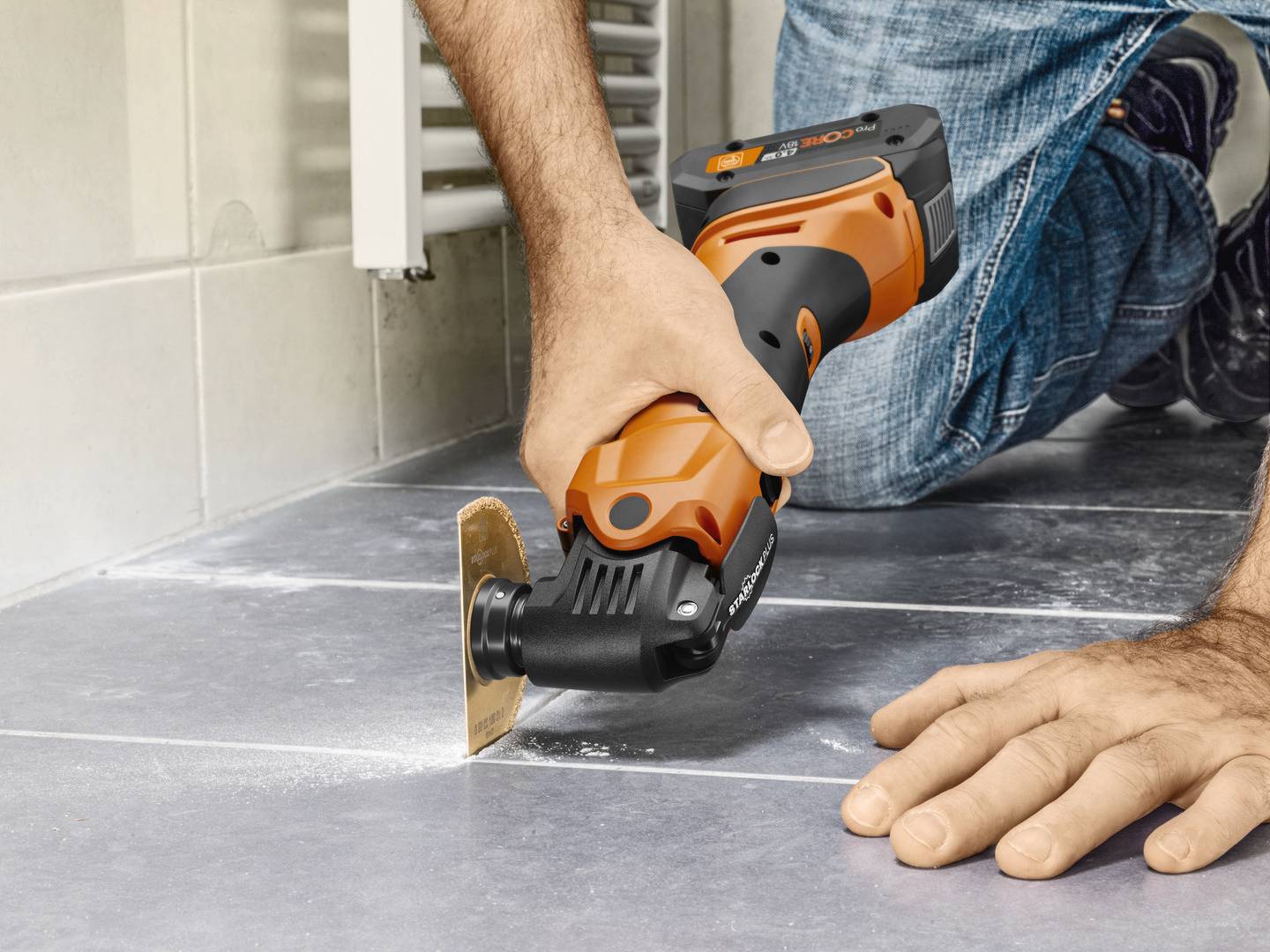 Een persoon snijdt vloertegels met een oscillerend gereedschap. Het apparaat heeft een oranje-zwart design. Focus op precies werken.