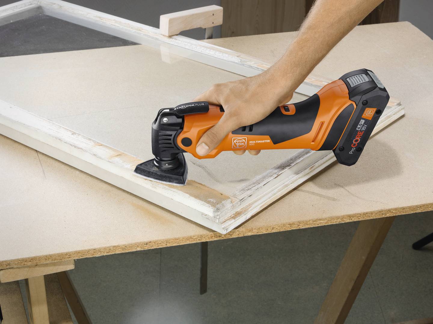 Een persoon schuurt met een oscillerend elektrisch gereedschap de rand van een houten frame op een tafel.