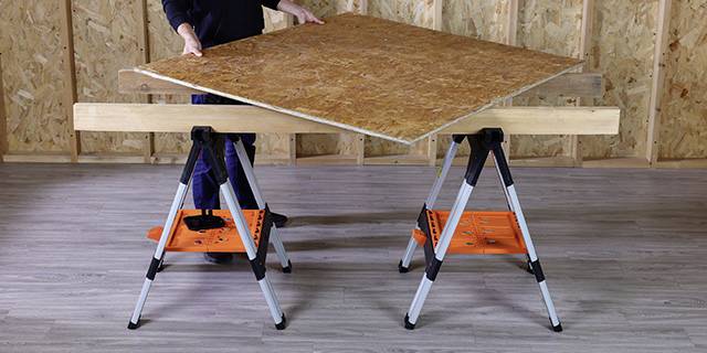 Een persoon legt een groot stuk multiplex op twee zwarte schragen in een werkplaats met houten wandbekleding.