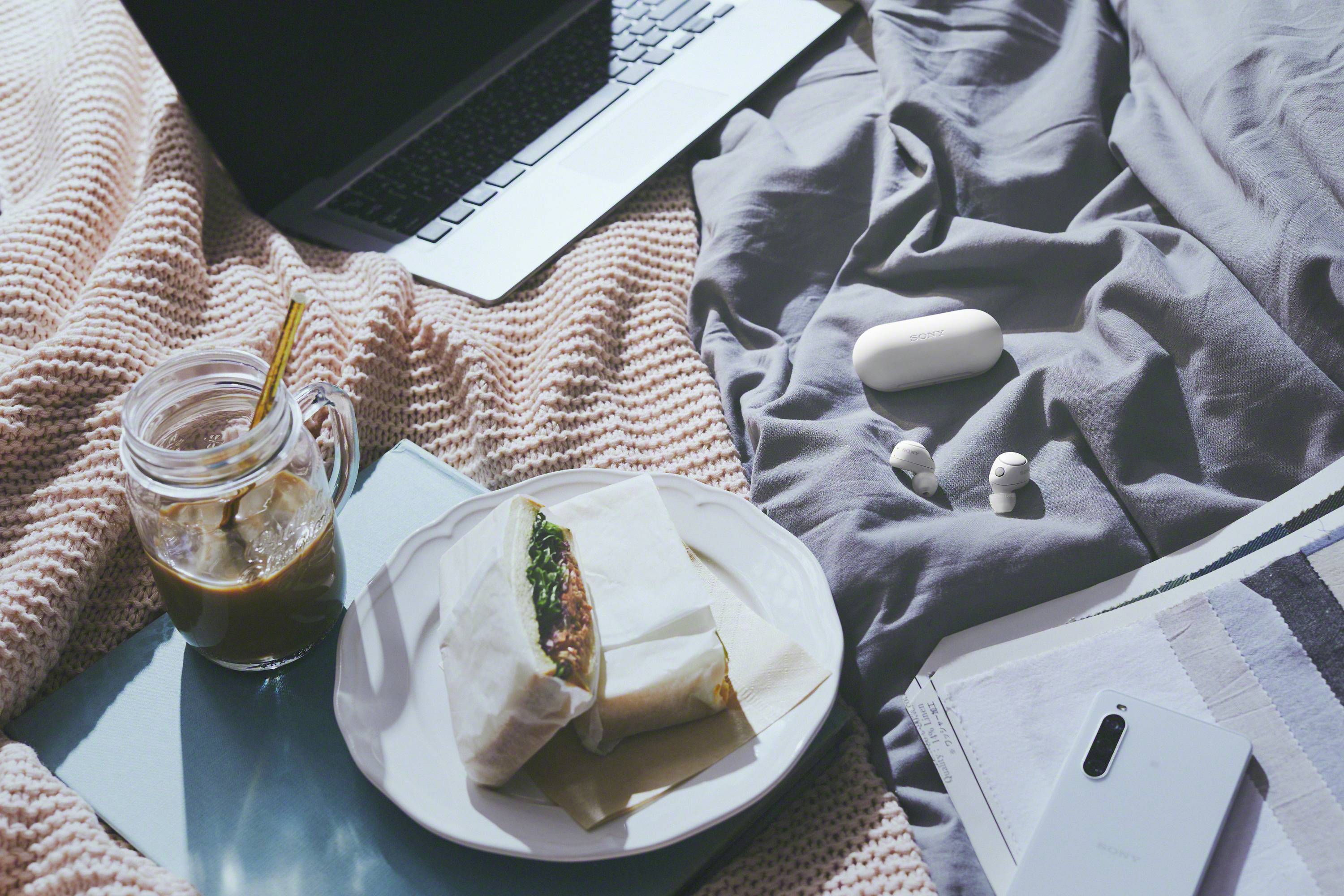 Op een bed liggen een laptop, een bord met een broodje, een kopje ijskoffie, draadloze oordopjes en een smartphone op een notitieboek.