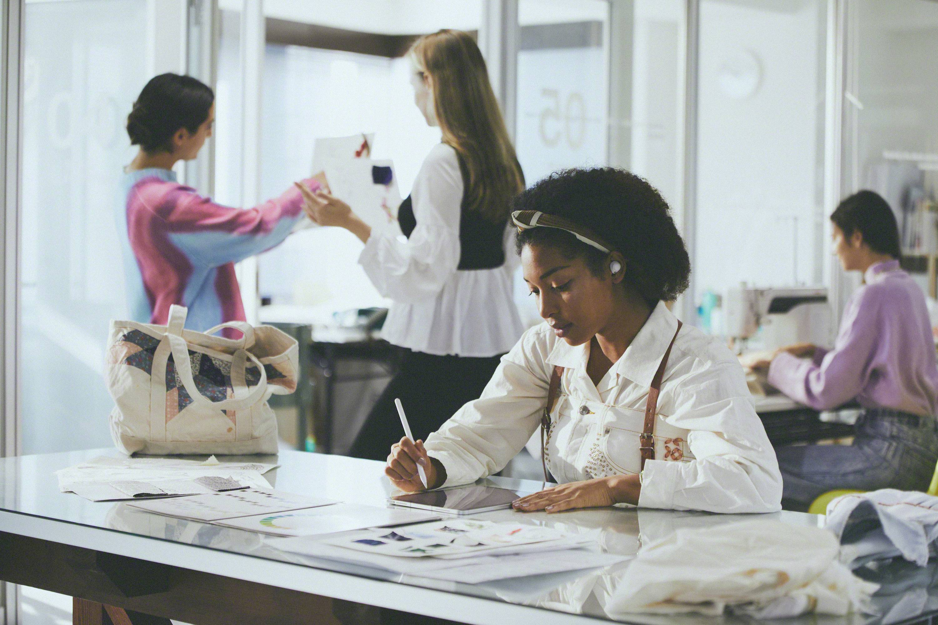 Een groep vrouwen werkt in een modern kantoor. Op de voorgrond tekent een vrouw aan een bureau, terwijl anderen op de achtergrond discussiëren en werken.