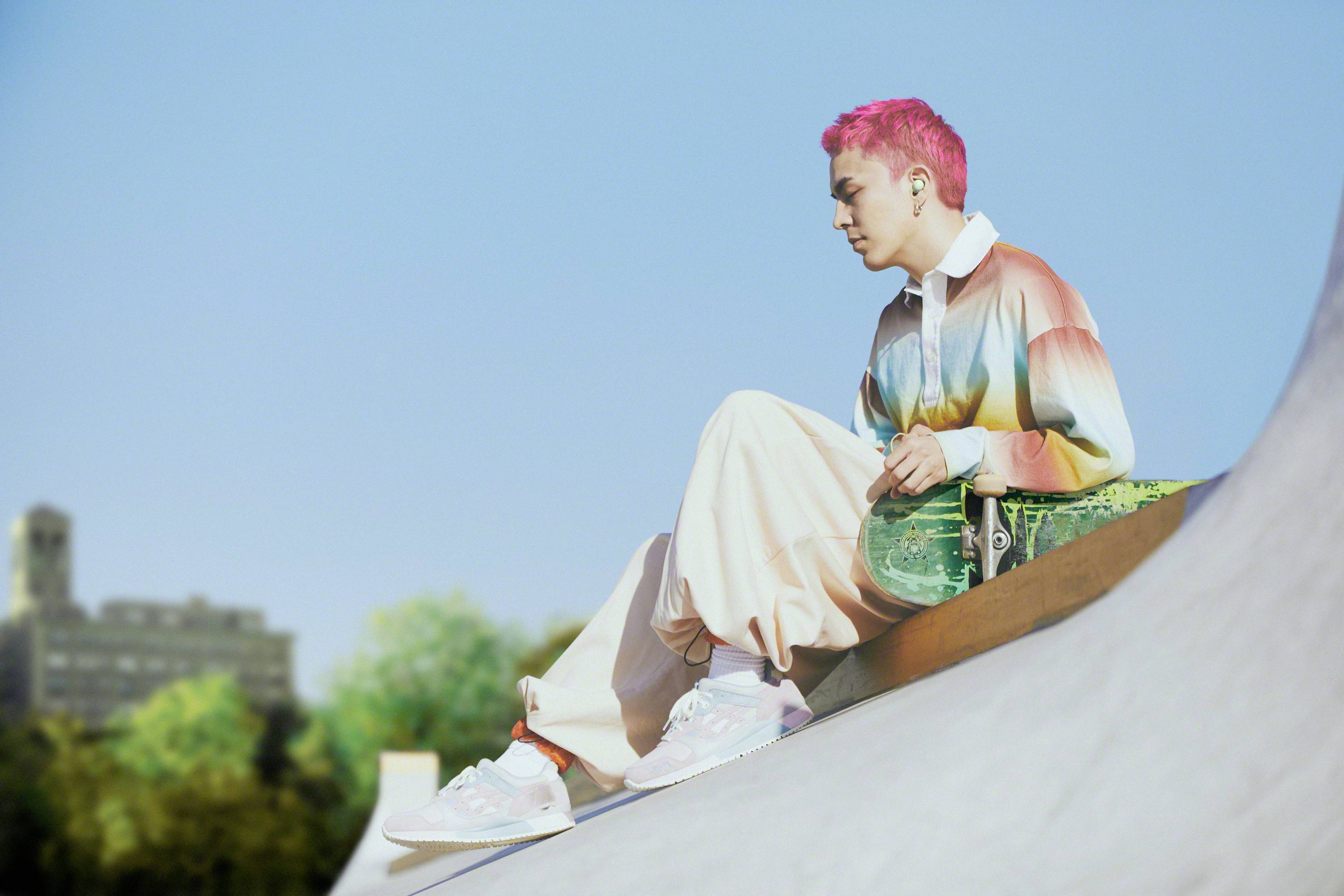 Een persoon met roze haar zit op een skateboard-ramp. Ze draagt kleurrijke kleding en houdt een skateboard vast, met gebouwen op de achtergrond.