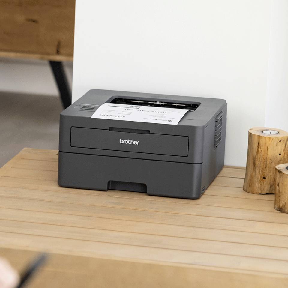 Een zwarte printer staat op een houten bureau. Ernaast liggen twee kleine houten planken. Op de achtergrond is een deel van een bruine tafel zichtbaar.