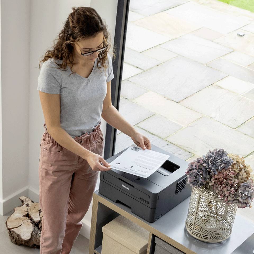 Een persoon drukt een document af van een printer in een lichte ruimte. Naast de printer staat een vaas met bloemen.