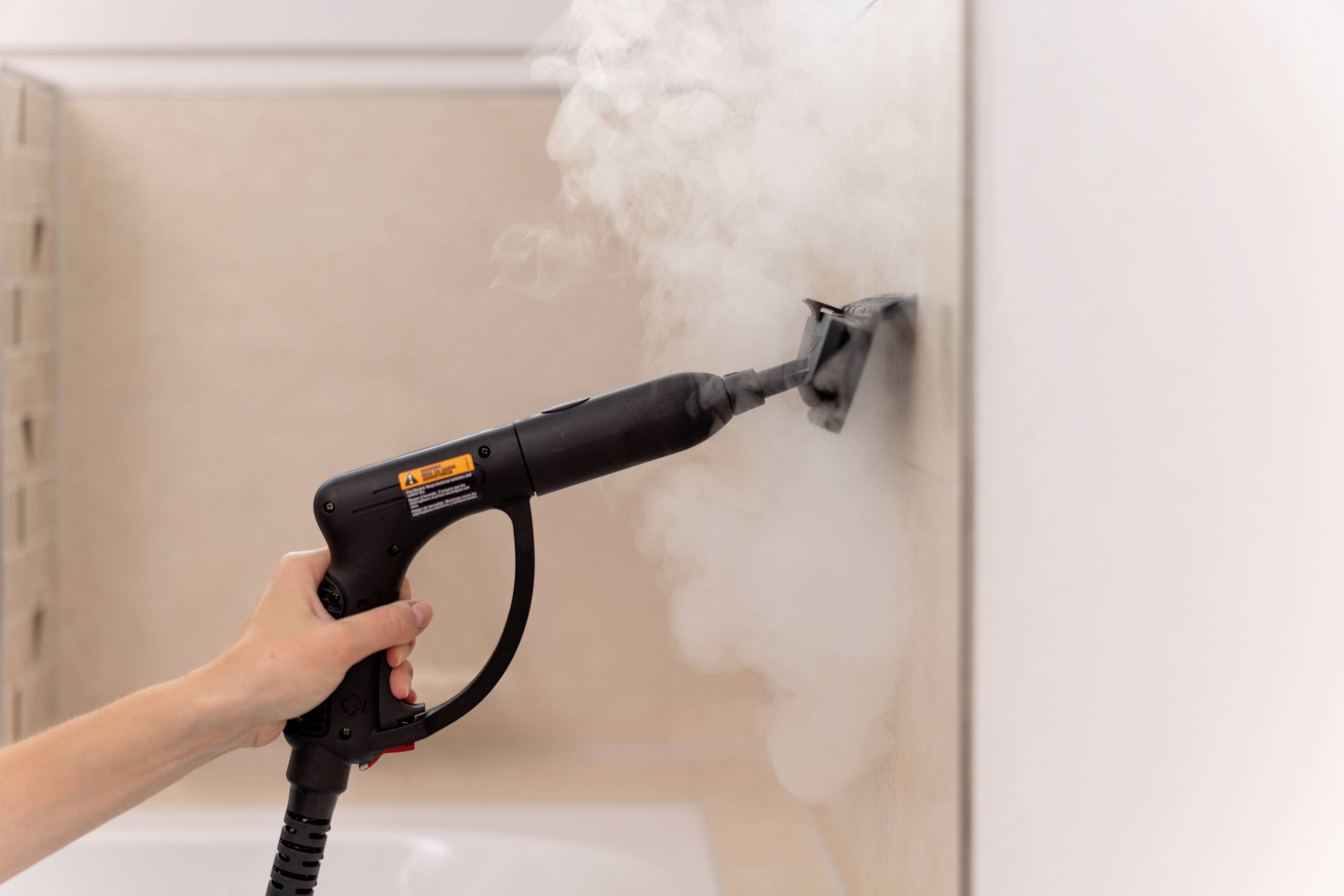Een hand houdt een stoomreiniger vast die stoom sproeit op een betegelde muur om deze schoon te maken.