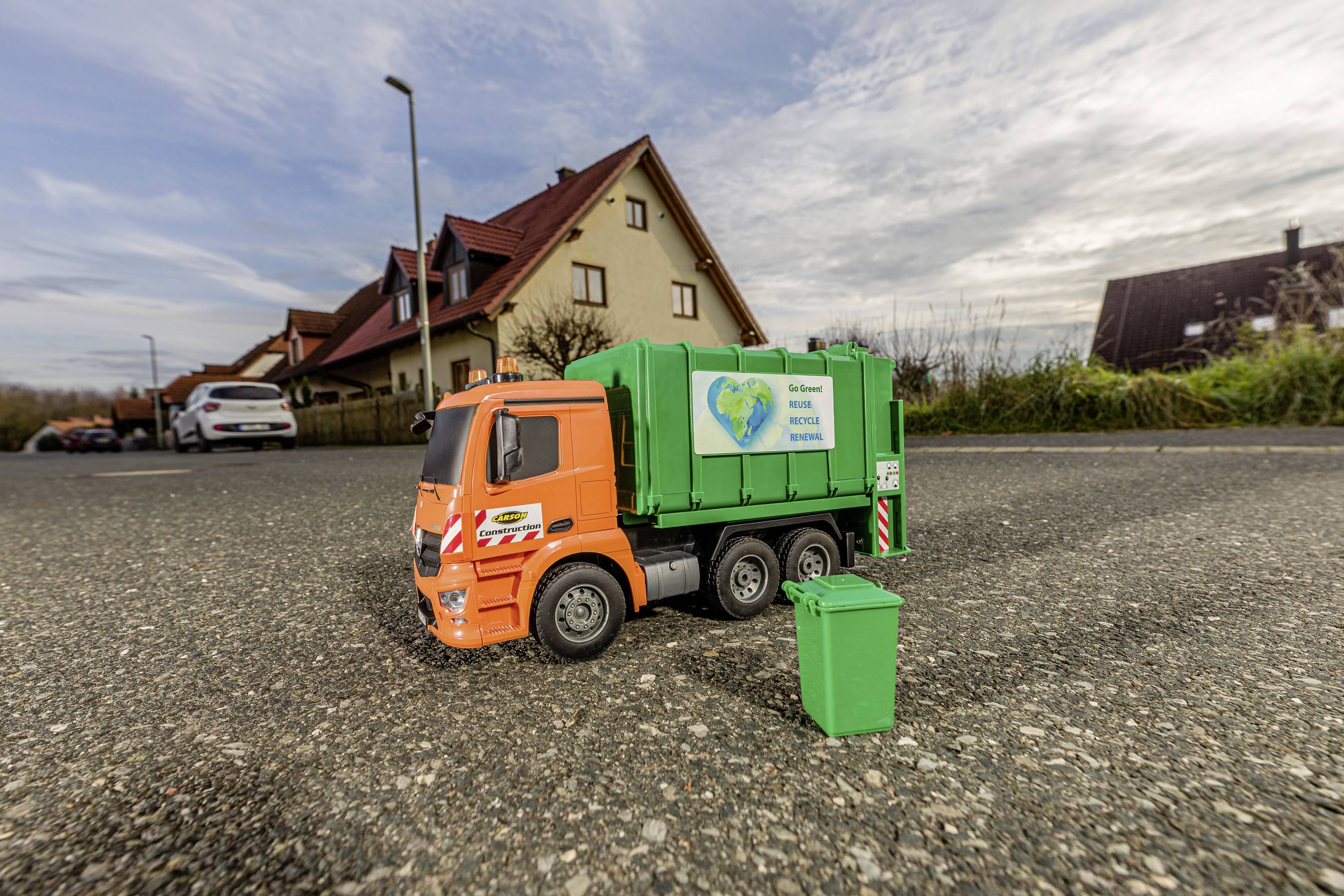 Speelgoed-vuilniswagen en vuilnisbak op een straat voor woningen. De vrachtwagen is oranje-groen met een milieuboodschap.