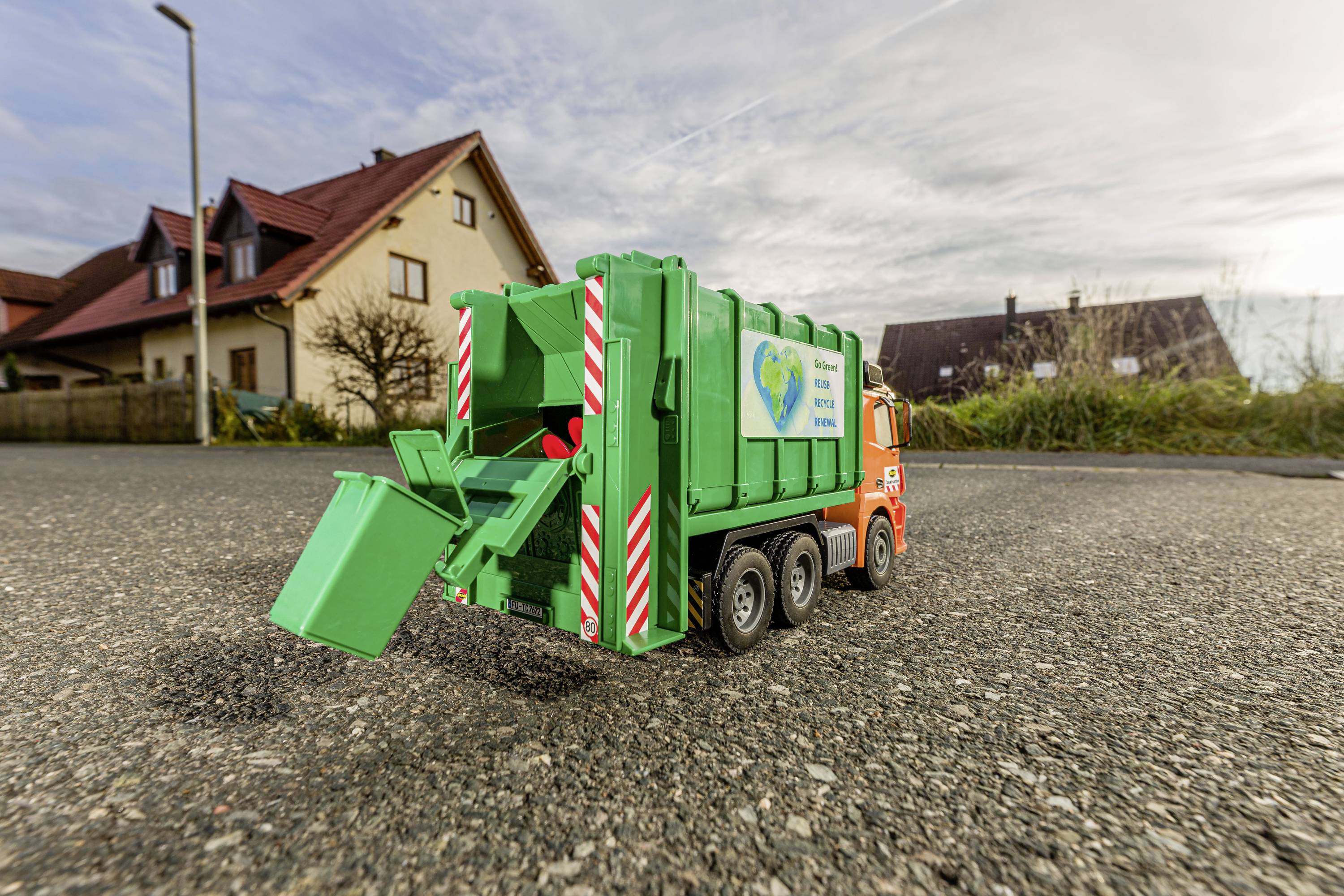 Een kleine speelgoed-vuilniswagen staat op een straat voor woonhuizen. Het groene voertuig heeft een open klep aan de achterkant.