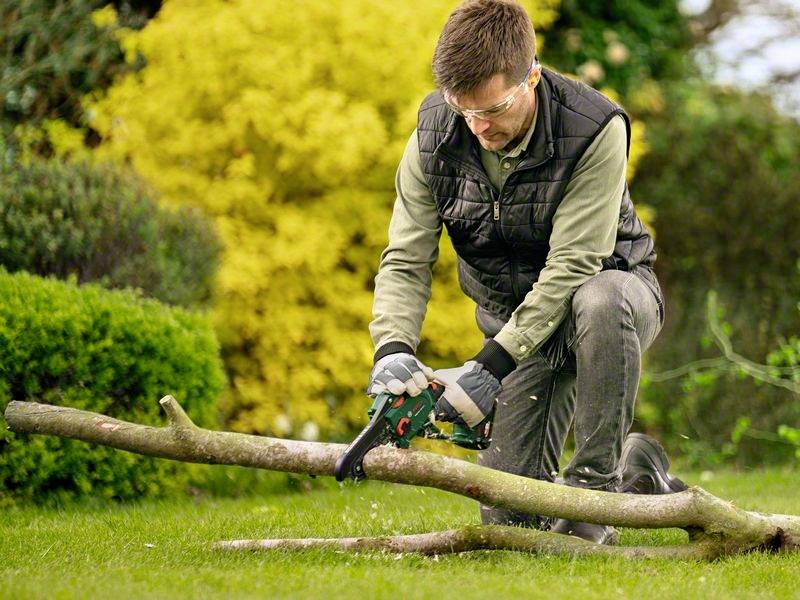 Een man met een veiligheidsbril en handschoenen knielt in de tuin en zaagt met een kettingzaag een boomstam door. Op de achtergrond zijn struiken zichtbaar.