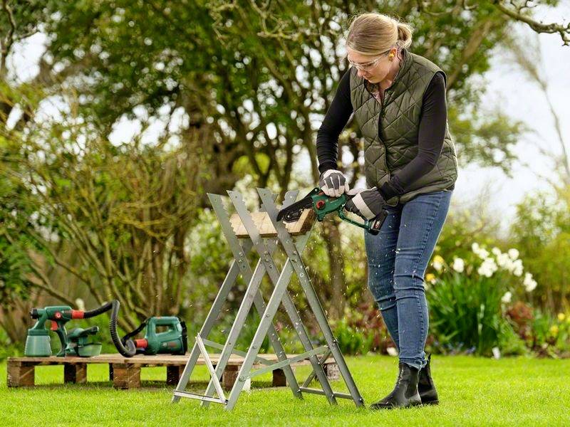 Een vrouw zaagt een houten plank op een zaagbok in de tuin. Op de achtergrond zijn struiken en tuingereedschap te zien.