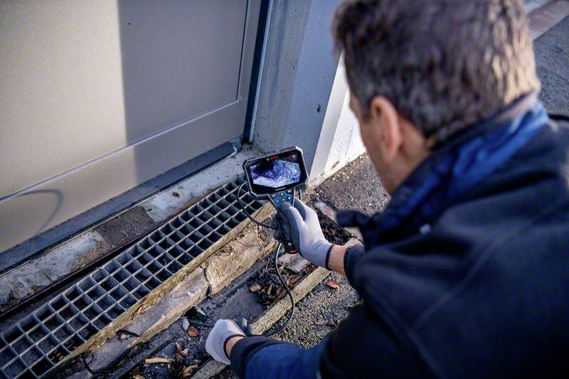 Een persoon inspecteert een rooster met een camera. Het scherm toont de binnenkant van het rooster. De nadruk ligt op het onderzoeksproces en de techniek.