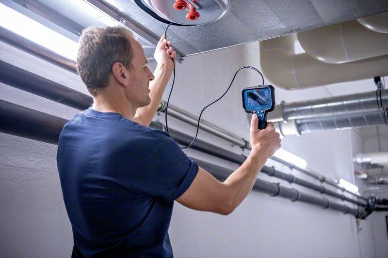 Een man gebruikt een inspectiegereedschap met een scherm om de binnenkant van een plafondventilatiebuis te inspecteren.