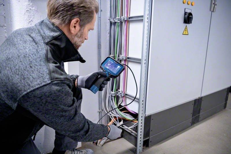 Een man inspecteert kabels in een schakelkast met een handapparaat. Veiligheidsuitrusting en kleurrijke kabels zijn zichtbaar.