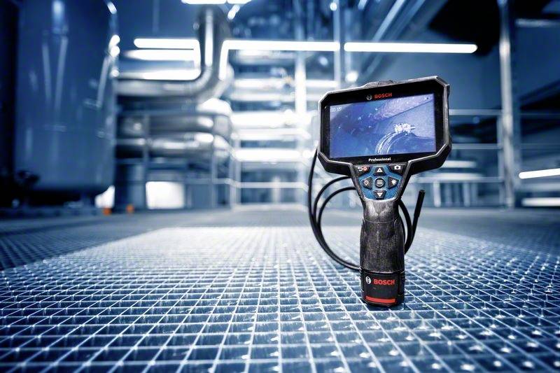 Een inspectieapparaat met een scherm en camera ligt op een metalen rooster in een industriële omgeving met buizen op de achtergrond.