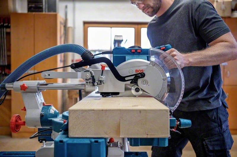 Een persoon gebruikt een tafelcirkelzaag in een werkplaats om nauwkeurige houtbewerking uit te voeren. Op de achtergrond zijn gereedschappen zichtbaar.