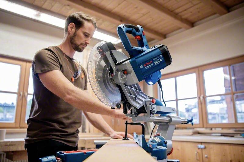 Een man werkt in een werkplaats met een blauwe afkortzaag. Op de achtergrond zijn houten rekken en grote ramen zichtbaar.