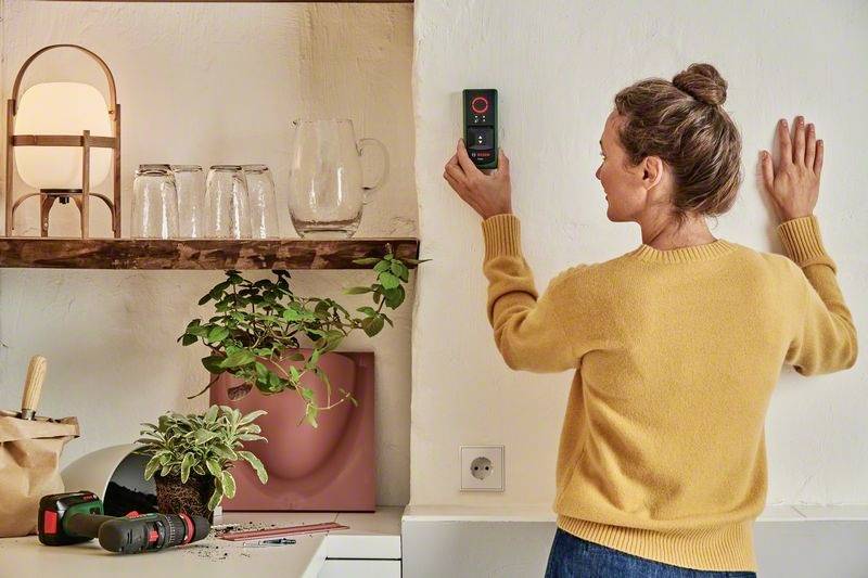 Een vrouw in een gele trui meet de muur met een digitaal meetapparaat. Op de achtergrond staan schappen met planten en glazen.