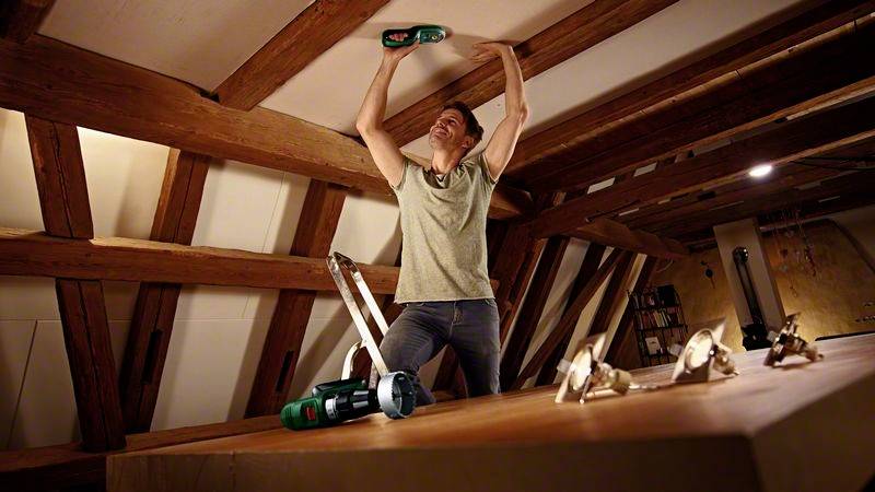 Een man bevestigt een plafondplaat in een zolder, omgeven door houten balken. Op tafel liggen gereedschappen.