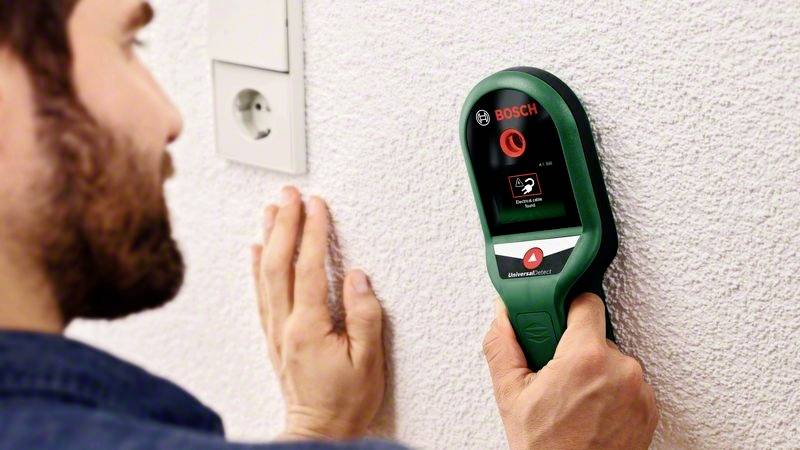 Een persoon houdt een groene Bosch-detector tegen de muur, waarop het scherm een detectiesignaal weergeeft.