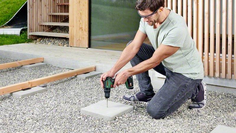 Een man boort met een accuboormachine een gat in een betonplaat op een grindondergrond voor een modern houten gebouw.