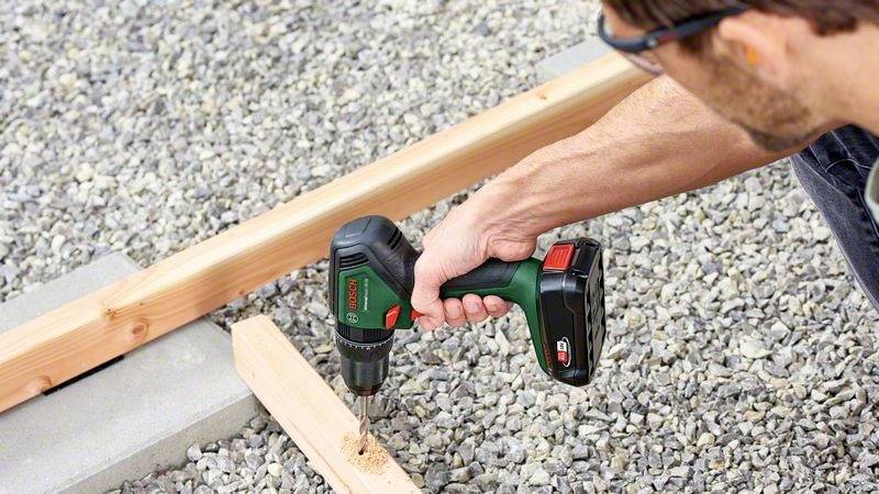 Een persoon boort met een accuboormachine een gat in een houten plank. Op de achtergrond zijn keien en een houten paal te zien.