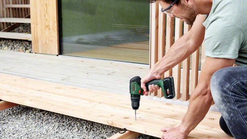 Een persoon schroeft houten planken aan een terras met een accuschroevendraaier. Ze draagt een veiligheidsbril en werkt buiten.