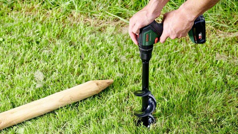 Een persoon gebruikt een accuboormachine met een aardboorboor om een gat in een gazon te boren. Naast hem ligt een houten paal op het gras.
