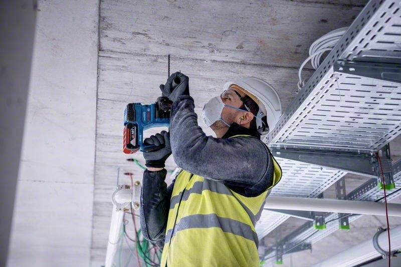 Een werknemer in beschermende kleding en helm boort in een plafond, omringd door kabels en buizen. Hij draagt een veiligheidsmasker.