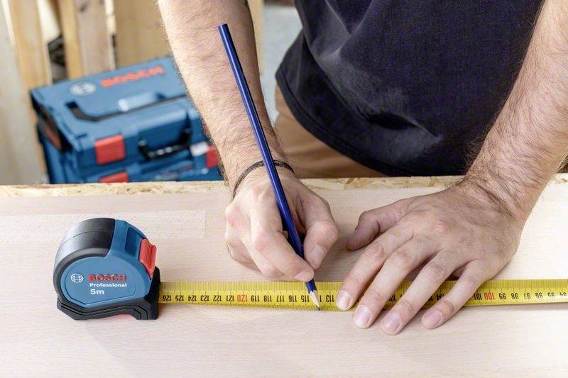 Een persoon meet een stuk hout met een meetlint en markeert het met een potlood. Op de achtergrond bevinden zich gereedschapskoffers.