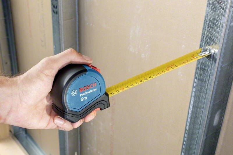 Een hand houdt een meetlint van "Bosch Professional" vast, meet de afstand tussen twee metalen frames, de schaal toont ongeveer 77 cm.