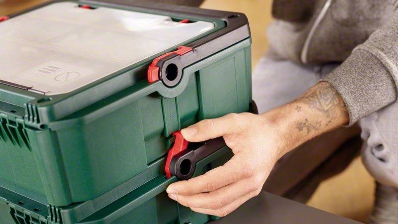 Een persoon stapelt twee groene gereedschapskisten door rode clips aan de zijkanten te vergrendelen.