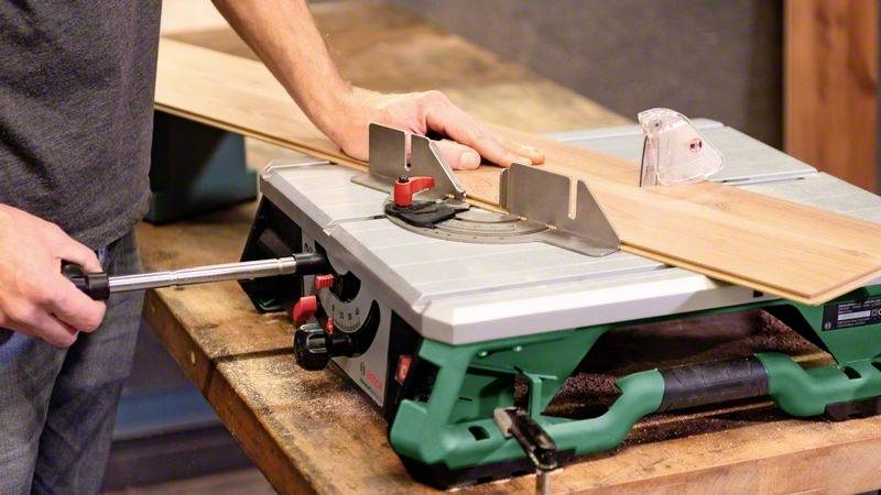 Een persoon zaagt een houten plank met een tafelzaag op een werkbank. Handen stellen de plank bij voor een rechte zaagsnede.