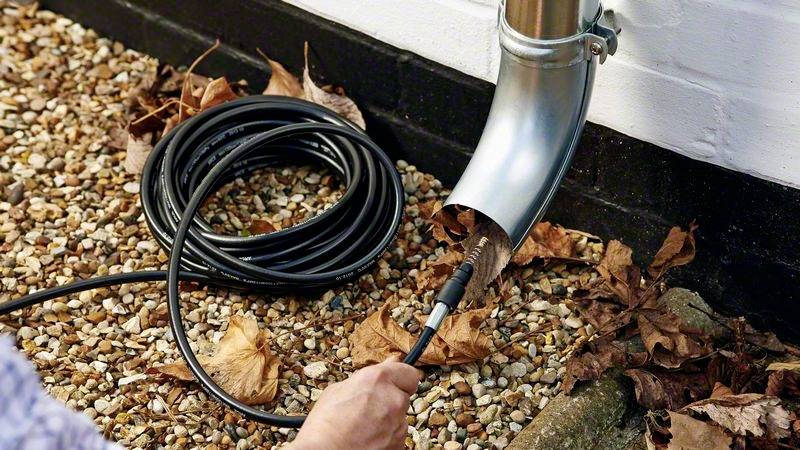 Een persoon reinigt een regenpijp met een buizenborstel. Herfstbladeren liggen op kiezels op de grond.
