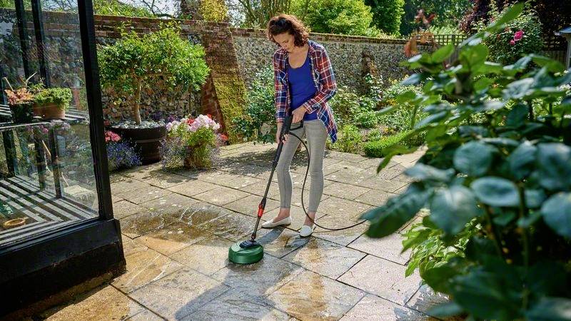 Een vrouw reinigt een terras met een hogedrukreiniger in een tuin. Op de achtergrond zijn planten en een stenen hek zichtbaar.