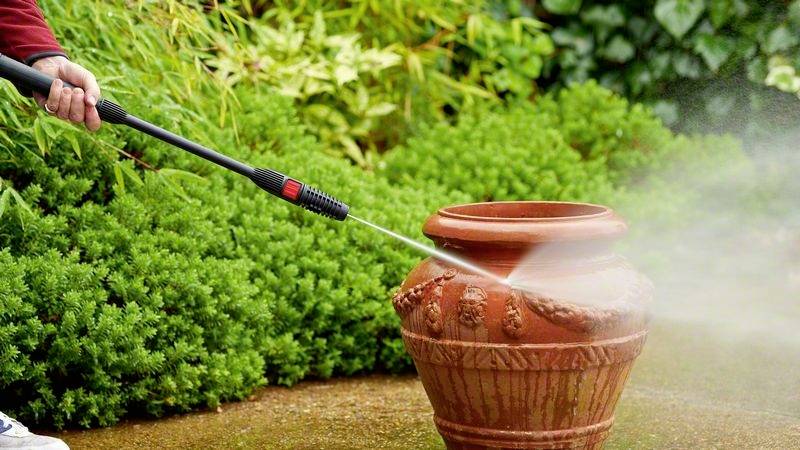 Een persoon reinigt een terracotta pot in de tuin met een hogedrukreiniger. Op de achtergrond zijn groene struiken te zien.