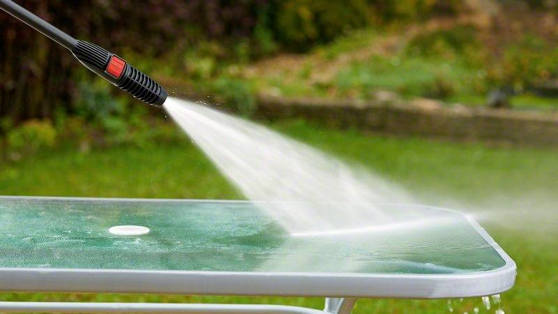 Een persoon reinigt een groene tafel buiten met een hogedrukreiniger. Water spat van de tafel af om vuil te verwijderen.