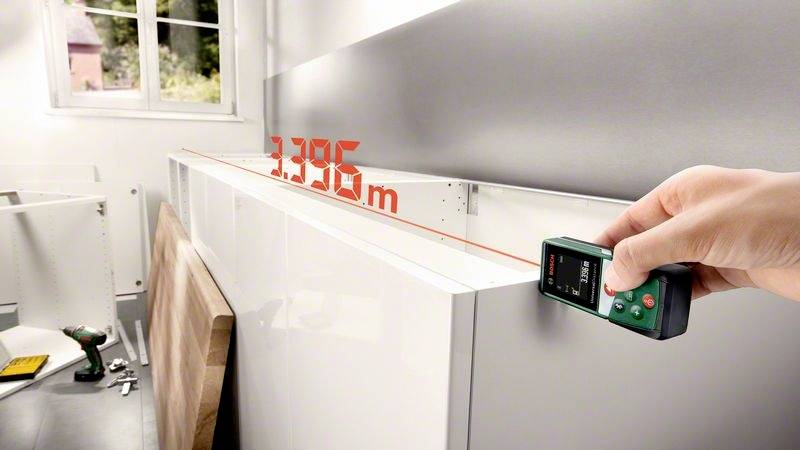Een persoon meet met een laserafstandsmeter de lengte van een keukenkast, waarbij '3,395 m' wordt weergegeven. Op de achtergrond is een raam zichtbaar.