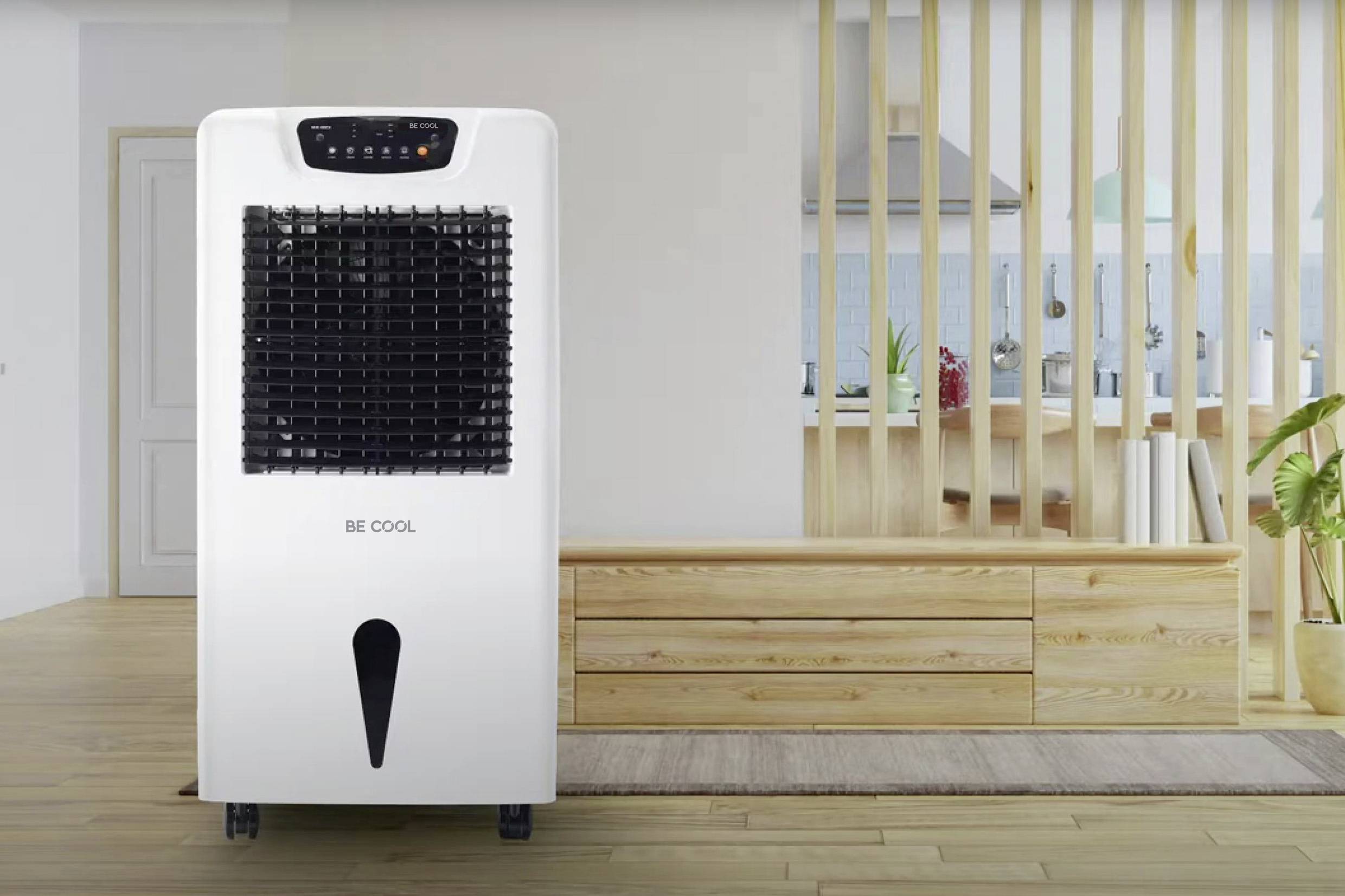 Het beeld toont een witte aircooler in een moderne woonkamer met houten vloeren en een open keuken op de achtergrond.