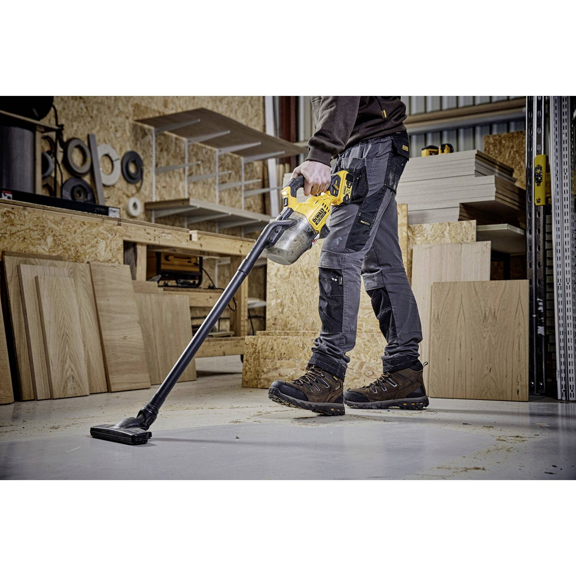Een werknemer gebruikt een gele stofzuiger om houtspaanders op te zuigen in een werkplaats met houten platen en planken.