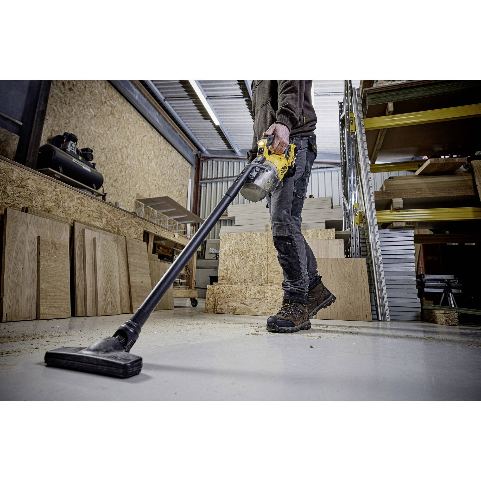 Een persoon zuigt de vloer van een houtworkshop; omringd door houten platen en rekken. Draagt werkkleding en houdt een gele industriële stofzuiger vast.