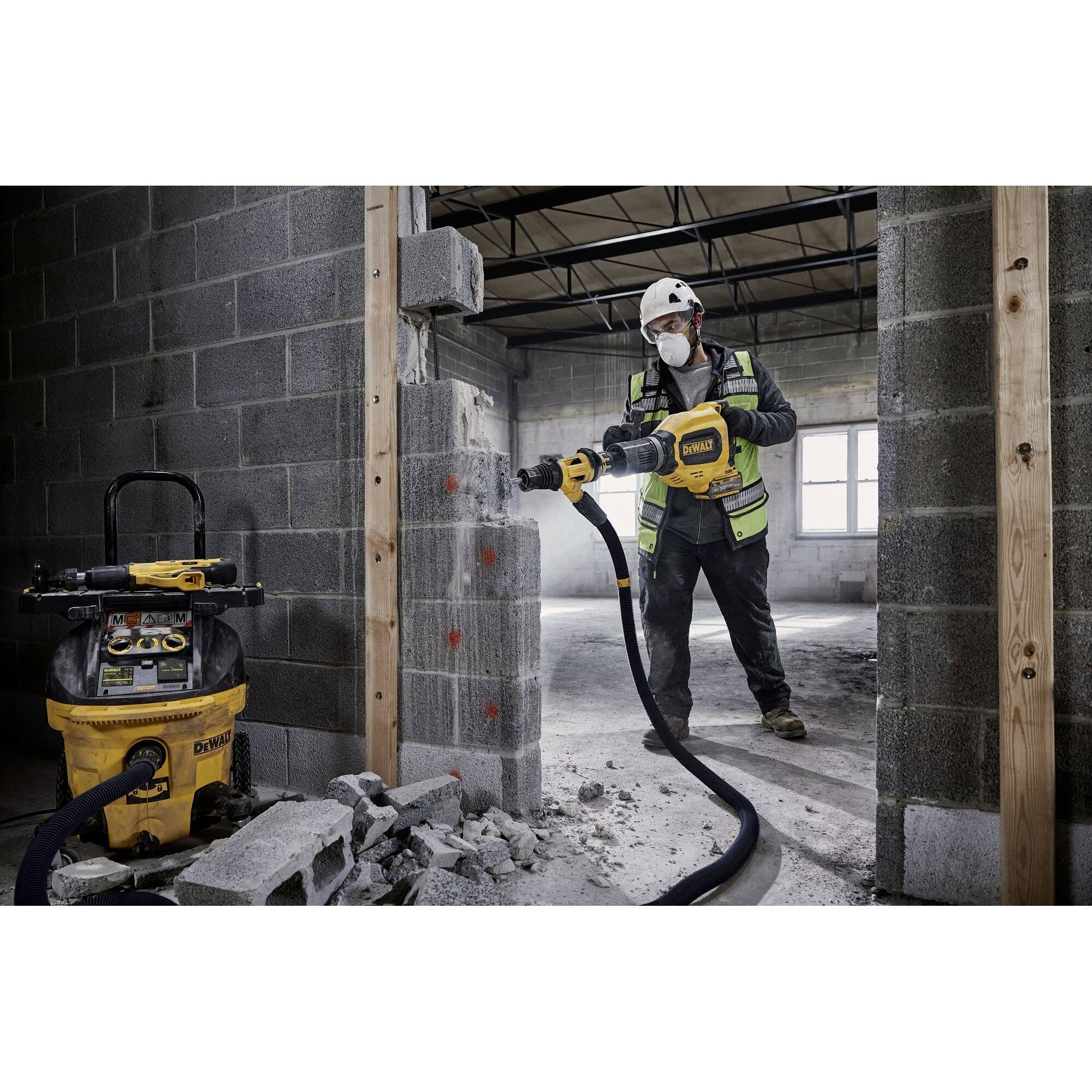 Een bouwvakker met veiligheidshelm en masker gebruikt een elektrisch sloopgereedschap om een betonnen muur in een casco-pand te doorbreken.