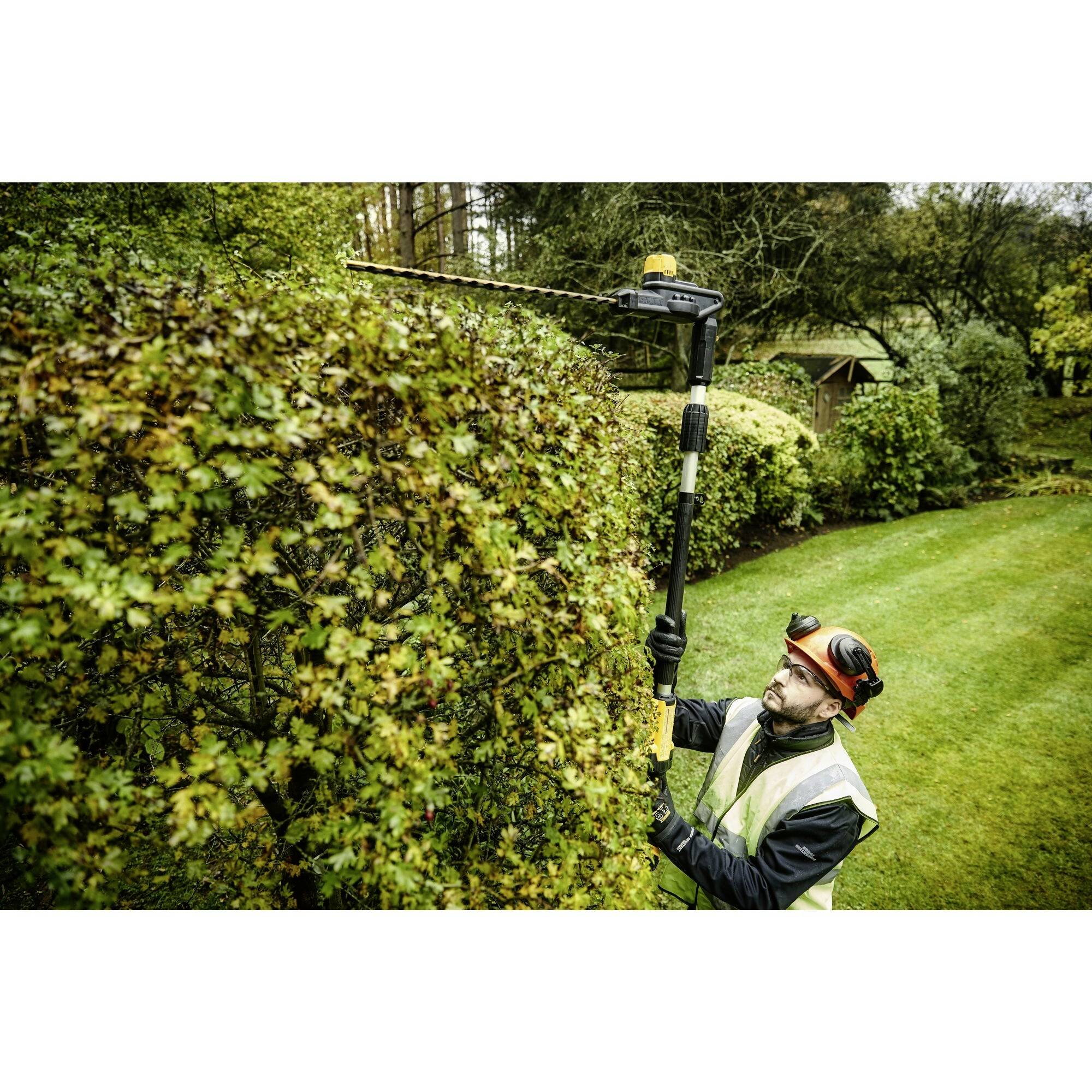 Een persoon in beschermende kleding knipt een hoge haag in een tuin met een elektrische heggenschaar op een telescopische steel.