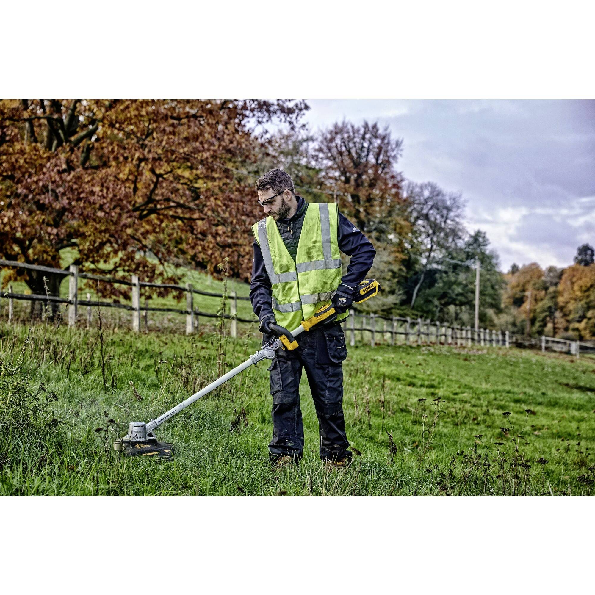 Een man maait gras met een trimmer op een weide. Hij draagt een gele veiligheidsvest. Op de achtergrond zijn bomen en een houten hek.