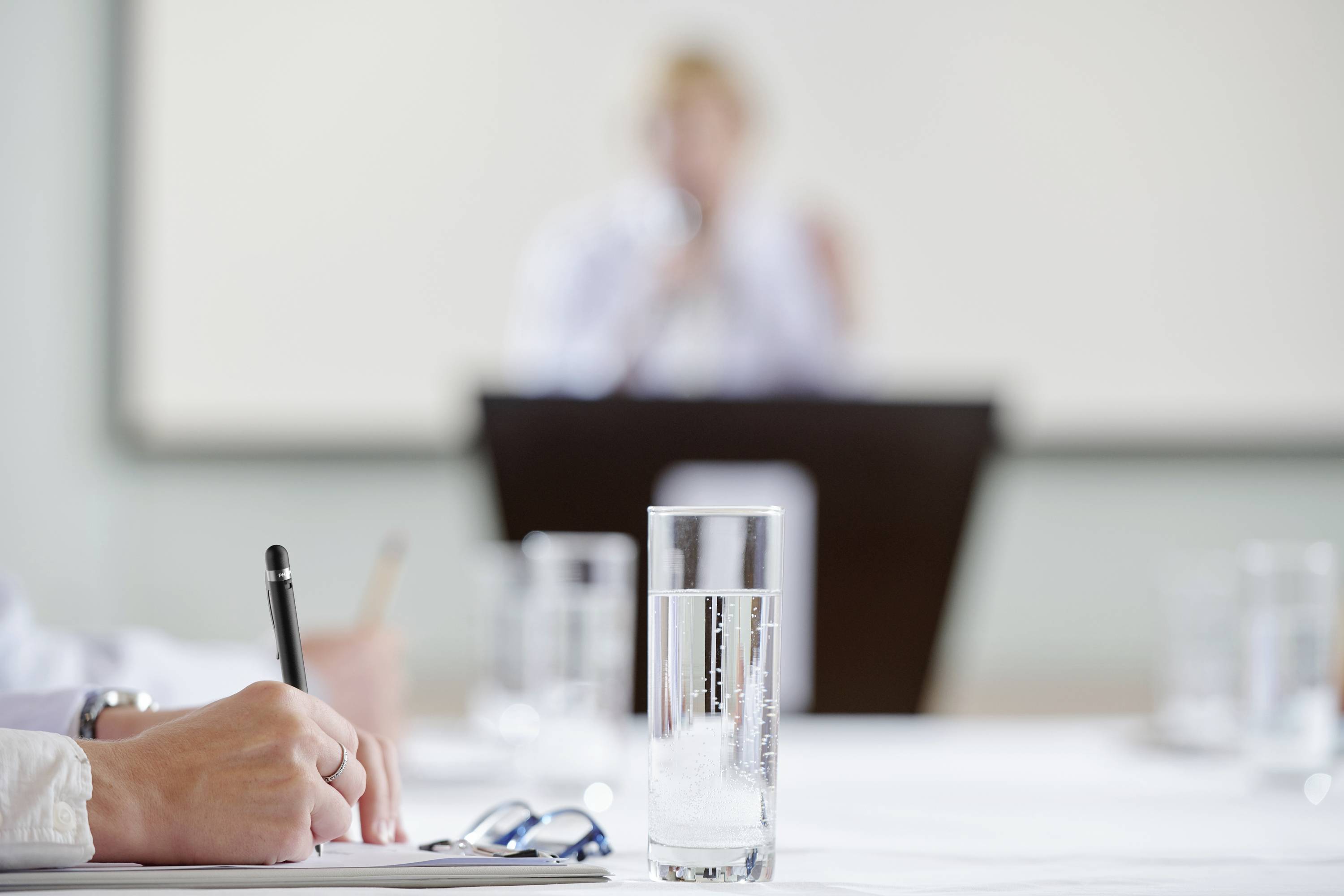 Een halfvol waterglas staat op de voorgrond van een tafel, met een wazig figuur aan een bureau op de achtergrond.