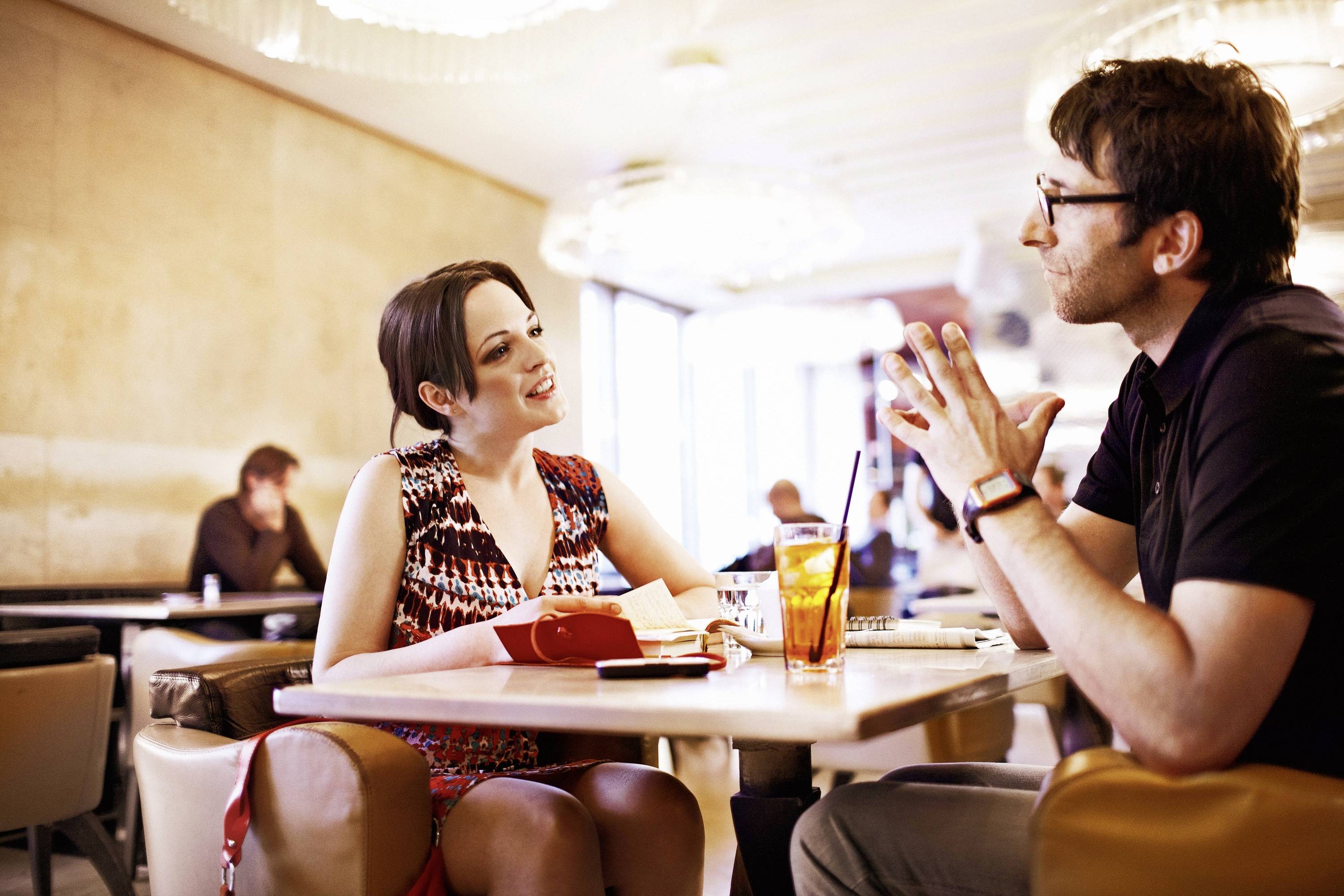 Een vrouw en een man zitten in een café en praten geanimeerd. Dranken staan op tafel, andere personen bevinden zich op de achtergrond.